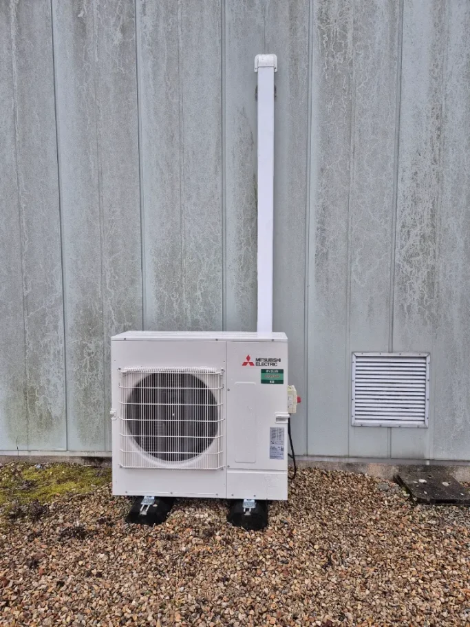 Installation d'un climatiseur extérieur Une unité extérieure de climatisation est posée sur un support, contre un mur en bardage métallique. Les conduites remontent verticalement.